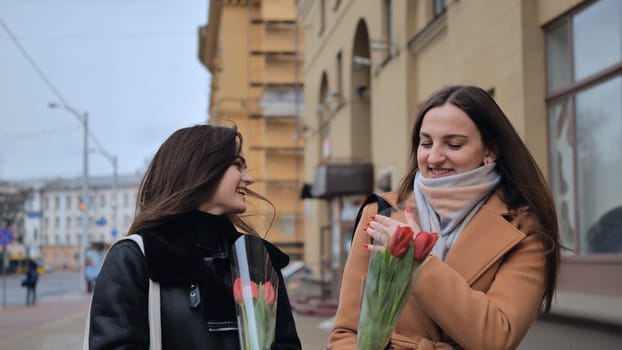 Two young girls, friends with flowers in their hands, walk along a city street on a cloudy spring day.