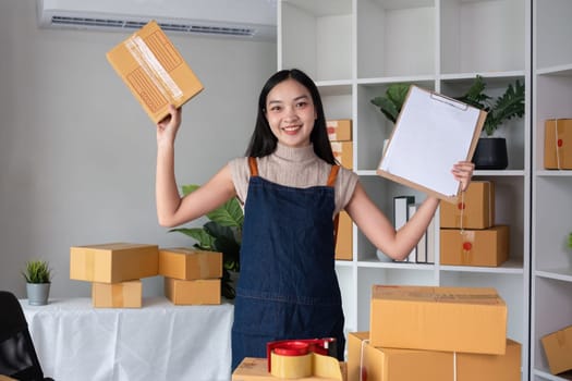Portrait of young female entrepreneur holding boxes of merchandise and clipboard listing customer shipping information..