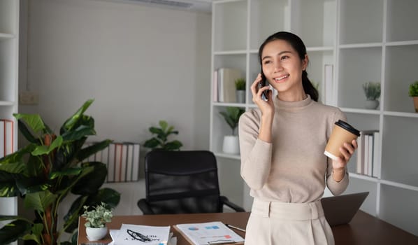 Asian businesswoman is talking on the phone in an online business meeting using a laptop in a modern home office decorated with lush green plants..