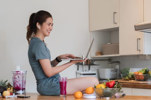 A woman is sitting at a kitchen counter with a laptop open and a bowl of fruit in front of her. She is smiling and she is enjoying her time