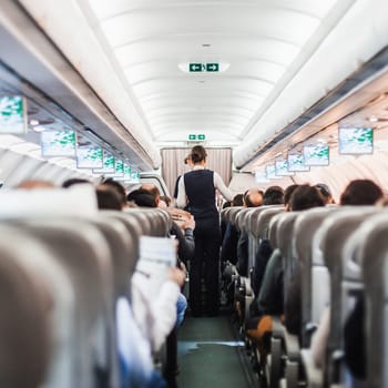 Interior of airplane with passengers on seats and stewardess in uniform walking the aisle, serving people. Commercial economy flight service concept