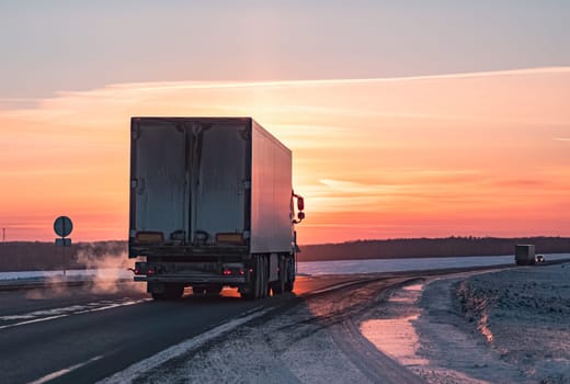 A semi truck cruises down a wintry highway as the sun sets on the horizon, casting a warm glow over the icy road and surrounding landscape.