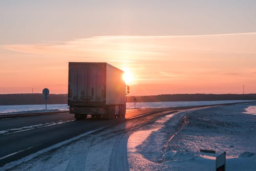 A semi truck cruises down a wintry highway as the sun sets on the horizon, casting a warm glow over the icy road and surrounding landscape.