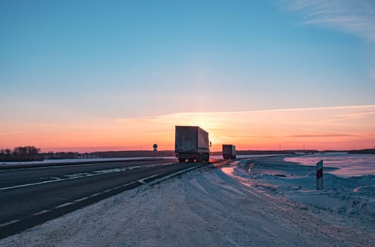 A semi truck cruises down a wintry highway as the sun sets on the horizon, casting a warm glow over the icy road and surrounding landscape.