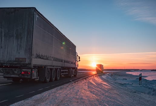 A semi truck cruises down a wintry highway as the sun sets on the horizon, casting a warm glow over the icy road and surrounding landscape.