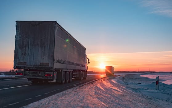 A semi truck cruises down a wintry highway as the sun sets on the horizon, casting a warm glow over the icy road and surrounding landscape.