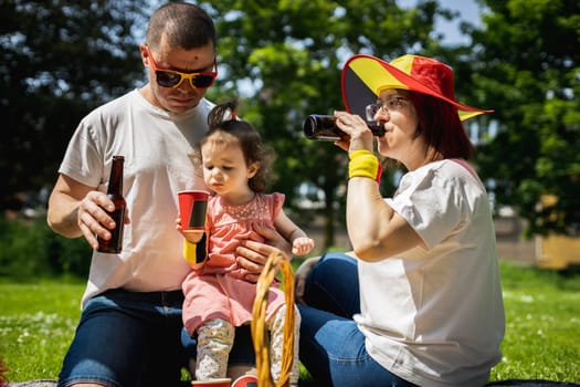 Portrait of beautiful and happy caucasian parents with little girls, drinks in their hands, celebrating Belgium day together at a picnic in a city park on a sunny summer day, close-up top view.