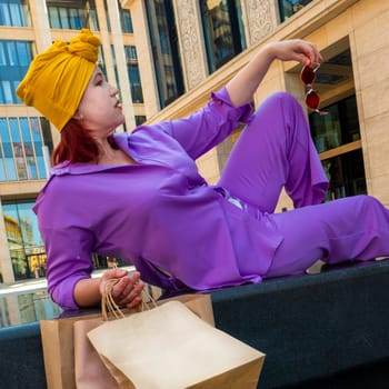 woman with craft shopping bags in the city. The concept of fashionable Consumerism and ecology. Adult Asian woman in a purple suit with red and yellow accents enjoys shopping. rest between purchases