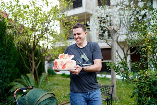 Smiling man with colorful birthday cake on a plate walking through the garden. High quality photo