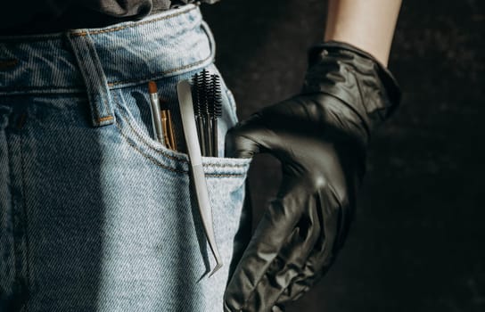 A set of brushes, tweezers and brushes for eyelash extensions stick out from the left pocket of blue jeans with a girl s hand in black gloves on a dark gray stone background, close-up side view. Dark style.