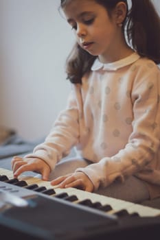 Beautiful little caucasian brunette girl with two ponytails plays the electric piano thoughtfully while sitting on the sofa in the room, top view close-up. Music education concept.