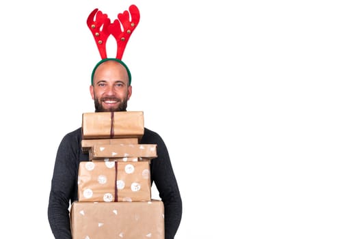 Portrait of young caucasian man with reindeer antlers looking at camera carries a pile of wrapped Christmas presents. Isolated on white background. Copy space. Holiday concept.
