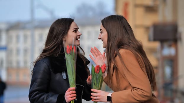 two happy girls, students, friends with flowers in their hands, laughing on a city street