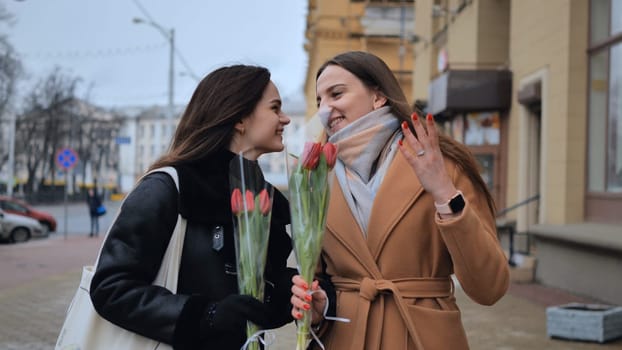 Two young girls, friends with flowers in their hands, walk along a city street on a cloudy spring day.