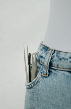 Two different tweezers and a round brush for eyelash extensions stick out from the right pocket of blue jeans, close-up side view from above.