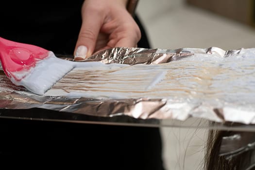 Hair coloring in a beauty salon. A master hairdresser-colorist dyes a client's brown hair blond. Apply lightening powder to hair onto foil using a pink brush. Close-up. Business concept.
