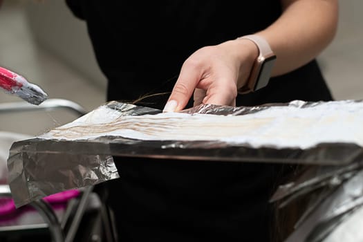 Hair coloring in a beauty salon. A master hairdresser-colorist dyes a client's brown hair blond. Apply lightening powder to hair onto foil using a pink brush. Close-up. Business concept.