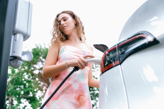 Eco-friendly conscious woman recharging modern electric vehicle from EV charging station. EV car technology utilized as alternative transportation for future sustainability. Synchronos