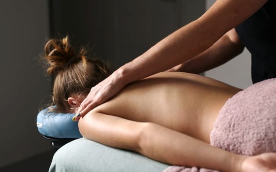 Professional Masseuse Massaging Back And Shoulder Blades Of Young Woman Lying On A Massage Table.