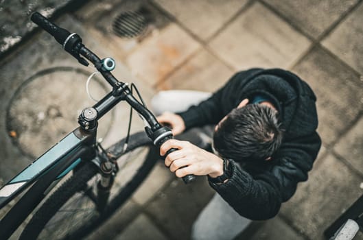 One young caucasian guy holds a bicycle handlebar with one hand and turns the handlebars with the other hand while squatting in the backyard of a house on a cloudy day, top view close-up with depth of field.