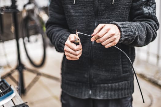 One young Caucasian unrecognizable guy in a dark gray cardigan is repairing his bicycle, warming up the brake wire with a fire using a lighter, standing in the backyard of a house on a cloudy day, close-up side view.