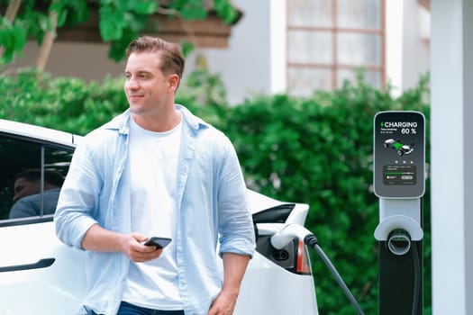 Modern eco-friendly man recharging electric vehicle from home EV charging station. Innovative EV technology utilization for tracking energy usage to optimize battery charging at home. Synchronos