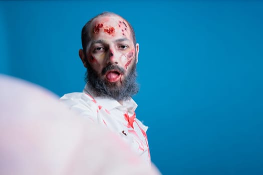 Portrait of brainless zombie isolated over blue studio background doing selfies with tongue out. Mindless undead creature covered in blood and wounds taking pictures with selfie camera