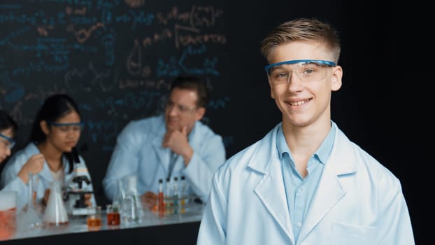 Smart student looking at camera while diverse group doing experiment. Attractive academic boy standing at blackboard with biological theory while people working by using chemical liquid. Edification.