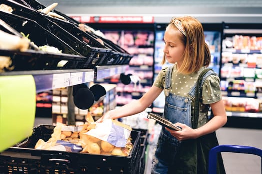 Pretty girl child buying vegetables with shopping list in supermarket and checking products with notes. Beautiful female preteen kid in grocery store