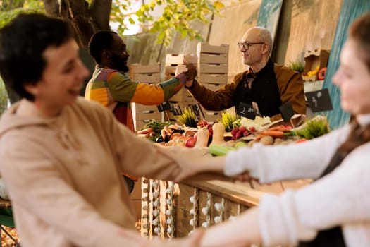 At farmers market stall, cheerful senior male seller gives black man a handshake and enjoys visit of a loyal customer. Joyful individuals having good moment at food fair with organic fresh produce.