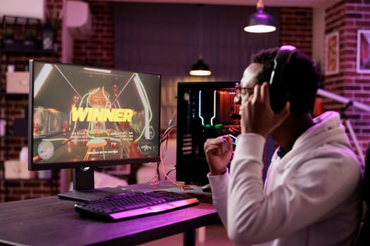 African american gamer excited about winning online multiplayer match against other players. Man seeing winner message on gaming PC computer display in neon lights living room