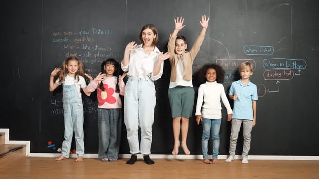 Multicultural student and teacher smiling and waving hand at blackboard with engineering code or prompt written. Smart children looking at camera and greeting while study in STEM classroom. Erudition.