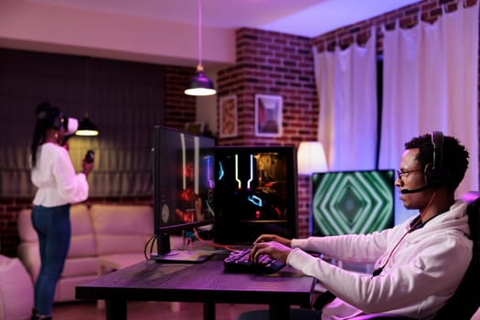 Gamer immersed in gaming session at computer desk while girlfriend in background uses virtual reality headset. African american couple playing games on gaming pc and vr system in rgb lights home