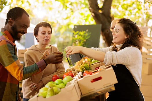 Happy multicultural couple tasting bio organic produce at farmers market, buying locally grown fruits and vegetables. Cheerful black man receiving slice of apple from friendly female vendor.