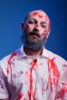 Portrait of actor portraying zombie character in horror movie with blood on face and clothes. Man dressed as undead creature for upcoming thriller film, wearing fake wounds makeup, studio background