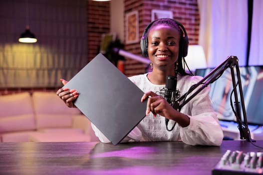 Portrait of african american tech content creator reviewing notebook in neon lights living room studio. Engaging content creator filming technology channel vlog for gadget aficionados, presenting info