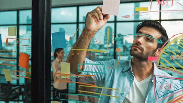 Professional businessman uses sticky notes at glass wall with graph to brainstorming idea while multicultural businesspeople discussing about marketing ideas at office with city view. Tracery