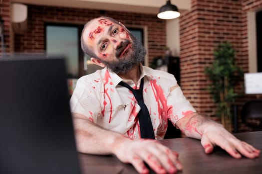 Portrait of overworked man with drowsy expression, limping on office desk chair, looking like zombie. Businessman covered in blood, looking exhausted after working too much, exploited by capitalism