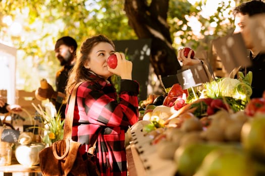 Standing in front of the farmers market stand, healthy customer enjoys the organic natural smell of apples. Before purchasing homegrown eco produce from the counter, a woman smells bio fruits.