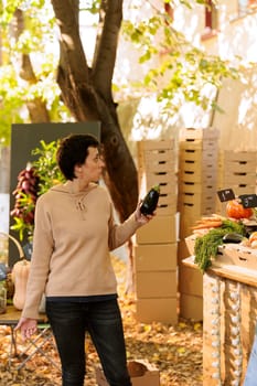 Young female customer with an eggplant in hand, purchasing locally grown colorful fresh natural produce. Woman shopper standing at farmers market booth, ready to buy freshly harvested veggie.