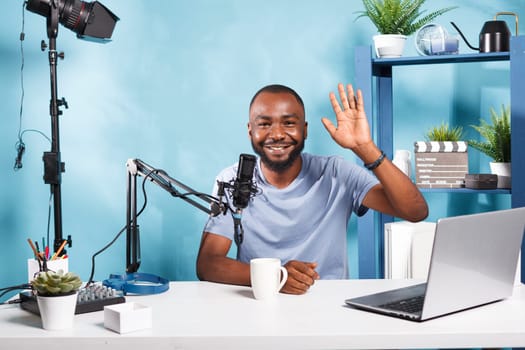 Smiling blogger recording video for online channel, waving hi to subscribers and looking at camera. Cheerful african american vlogger streaming live while working in home studio portrait