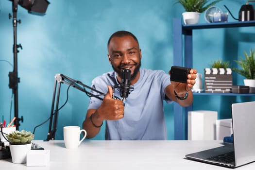 Smiling videographer recording camera gear positive review and showing thumb up gesture. Blogger live streaming and talking about photography equipment and holding accumulator