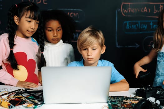 In classroom student in STEM class learning about coding robotics car. Smart children watching at laptop and thinking seriously but a child in bib play with a remote car and look at camera. Erudition.