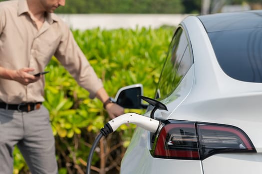 Young man use smartphone to pay for electricity at public EV car charging station green city park. Modern environmental and sustainable urban lifestyle with EV vehicle. Expedient