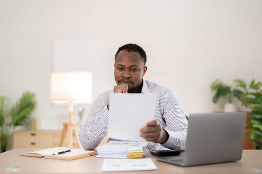 African American businessman feeling stressed while working on laptop, taking notes on documents in office and preparing work report..