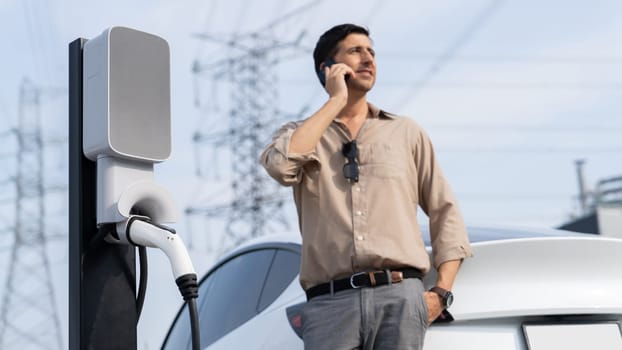 Man talking on the phone while recharge EV car battery at charging station connected to power grid tower electrical as electrical industry for eco friendly car utilization.Expedient