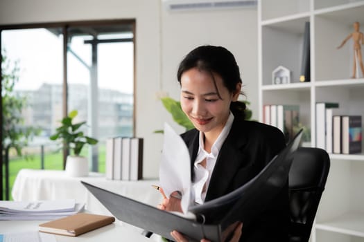 Focused Asian female accountant doing paperwork in office with plan documents on desk.