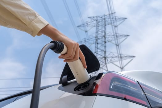 Closeup woman recharge EV electric car battery at charging station connected to electrical power grid tower on sky background as electrical industry for eco friendly vehicle utilization. Expedient
