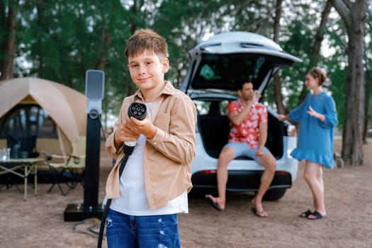 Little boy holding EV charger and point at camera with his family sitting on the trunk in background. Road trip travel with alternative energy charging station for eco-friendly car concept. Perpetual