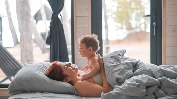 Red-haired Caucasian woman lies in bed with her baby son in a country house with panoramic windows
